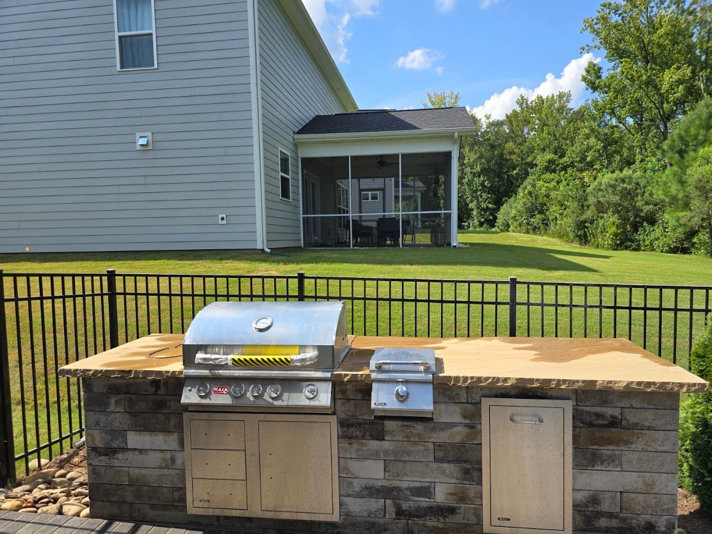 Outdoor kitchen with a grill and side burner, set in a backyard near a screened-in porch and fence.