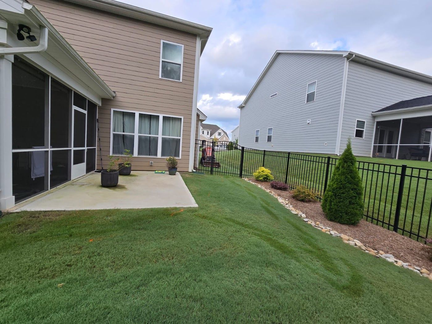 Lawn with two houses; one brick, one gray. Black fence and green grass. Cloudy sky.