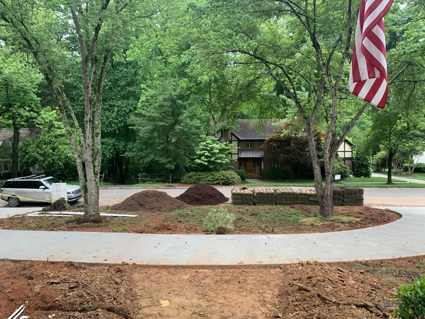 Street with landscaped island, mulch piles, trees, parked vehicle, and American flag.
