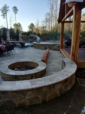Stone patio with fire pit, hot tub, and wooden structure in a backyard setting.