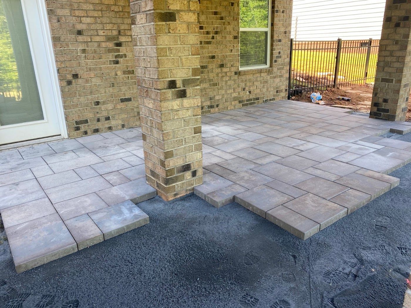 Patio construction in progress; pavers being laid on gravel base next to a brick pillar and house.