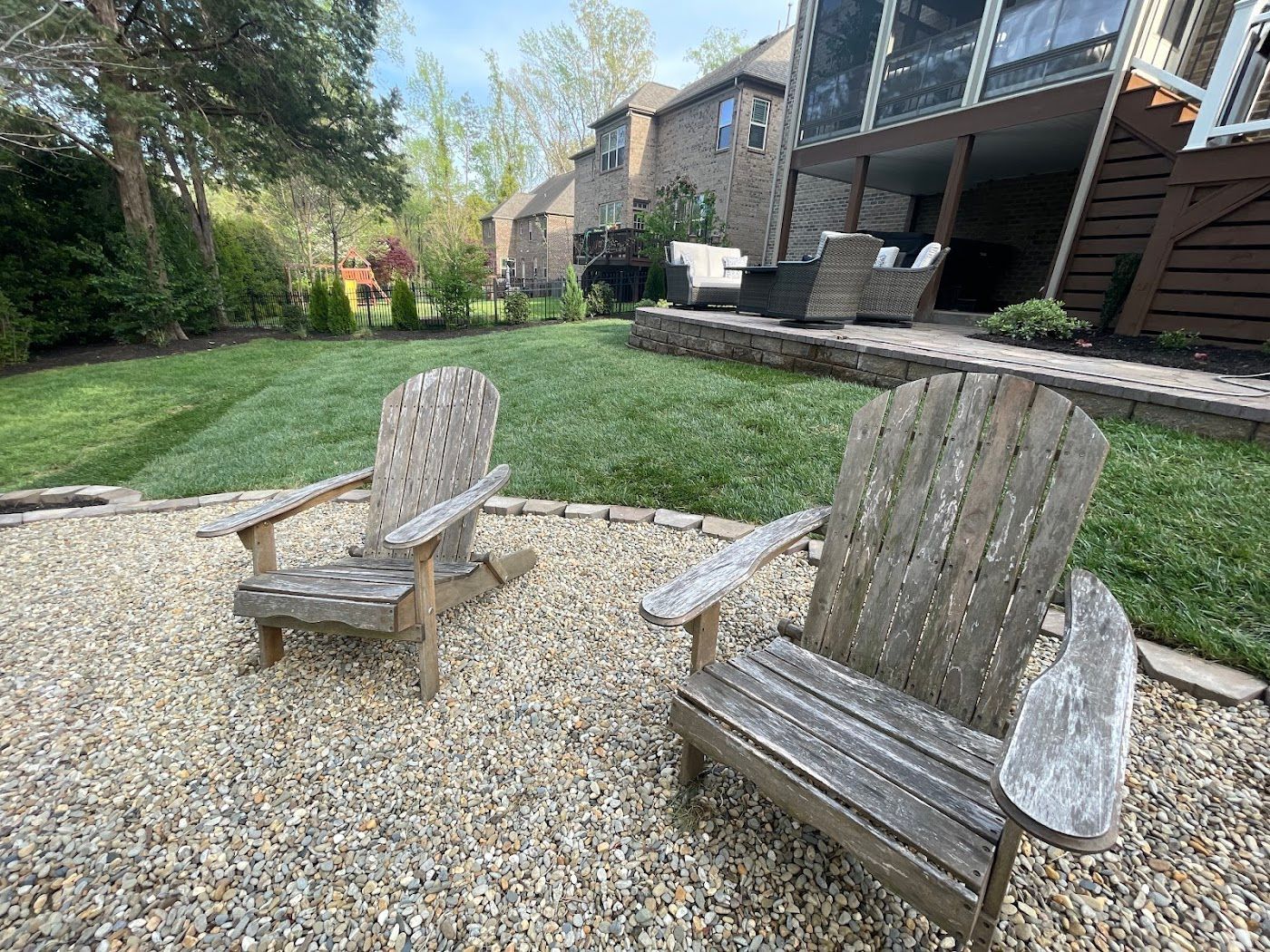 Two weathered wooden Adirondack chairs sit on gravel, facing a grassy backyard and a multi-story brick house.