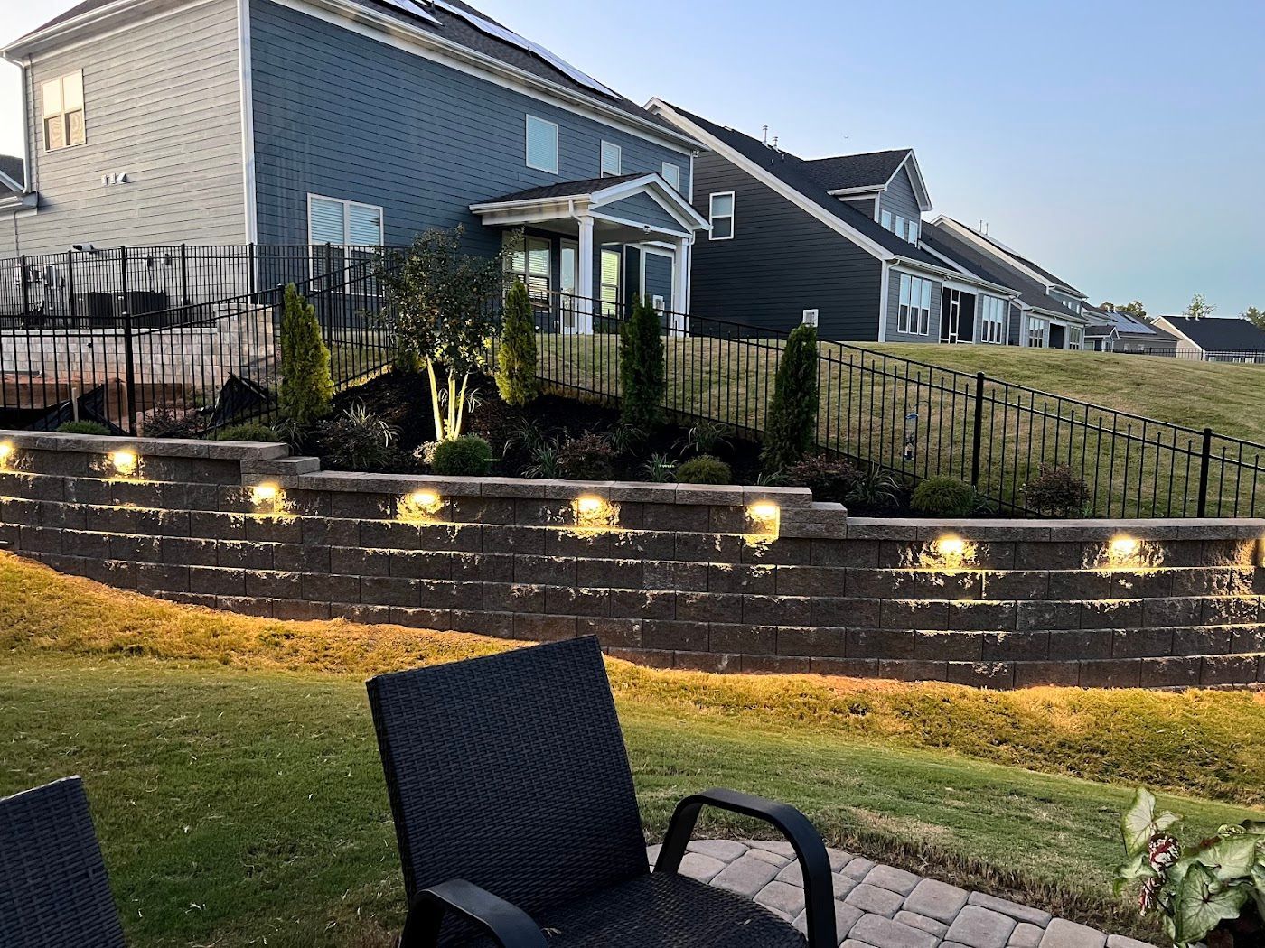 Backyard with illuminated retaining wall, shrubbery, and houses.