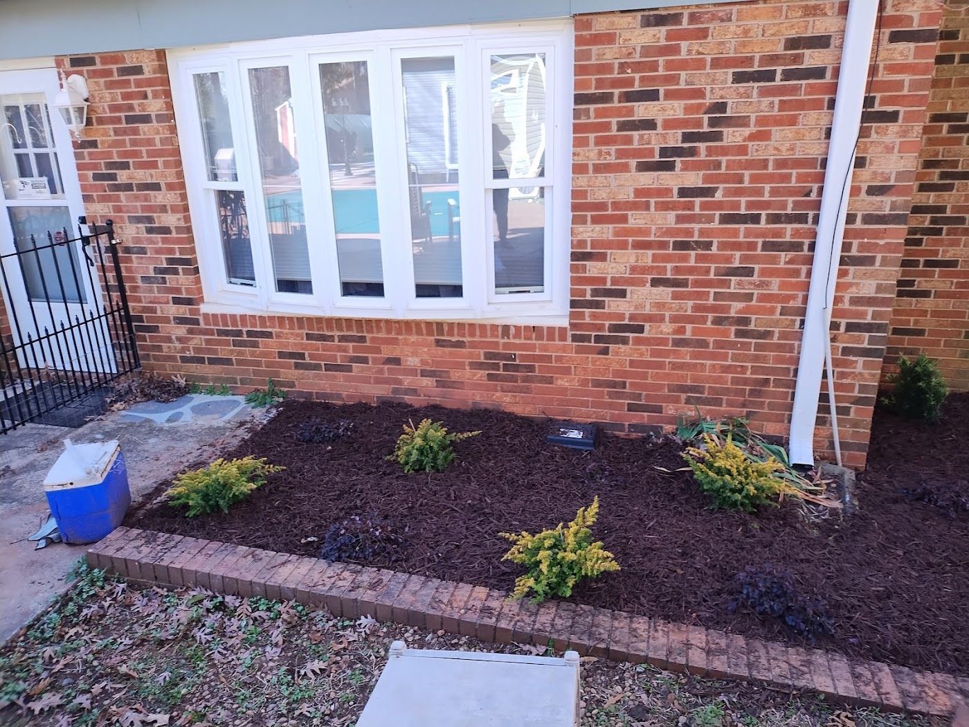 Brick house with a flower bed of mulch and green bushes. A window is in the center.