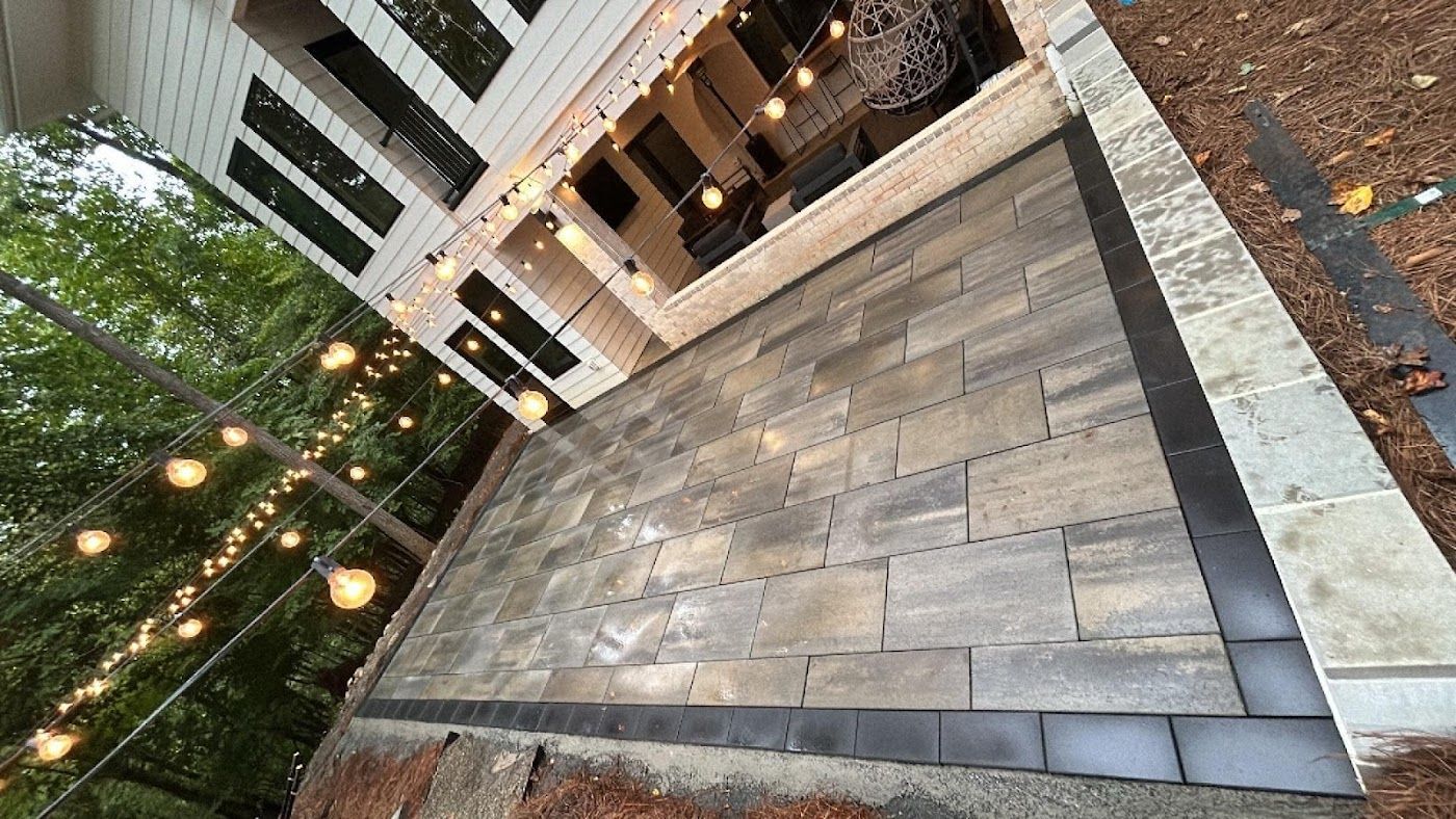 Patio with gray pavers next to a white house, illuminated by string lights.