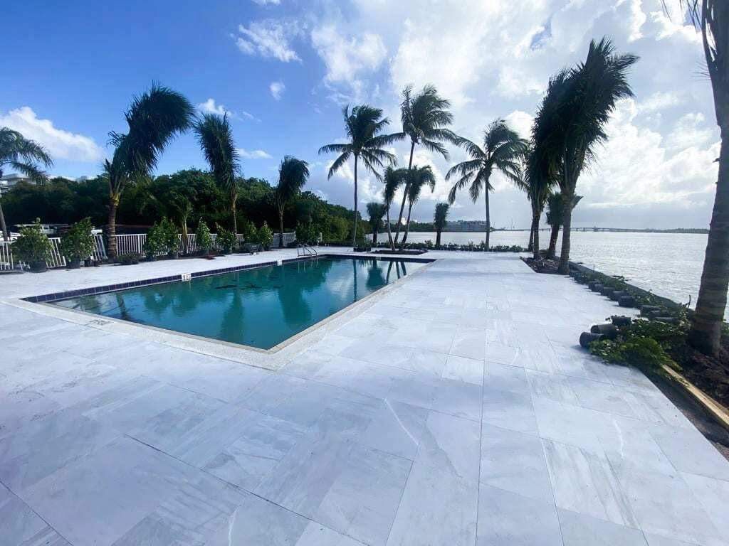 Pool with white stone patio, palm trees, and water view under a cloudy sky.