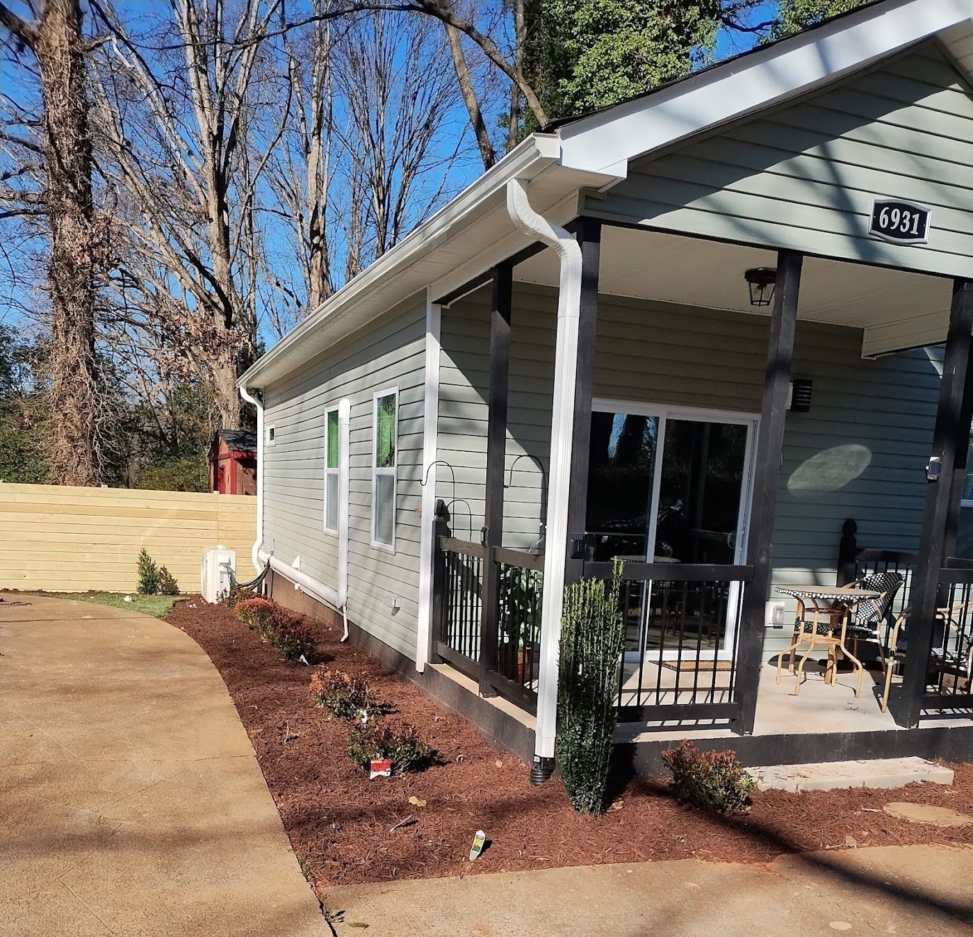 Small house with light green siding, black porch, and brown mulch landscaping.
