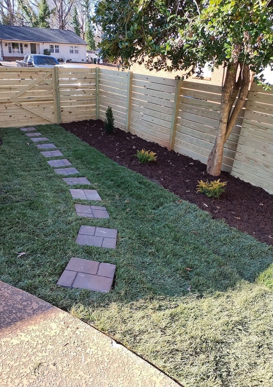 Lawn with stepping stones, mulch bed, and horizontal wooden fence beside a tree.