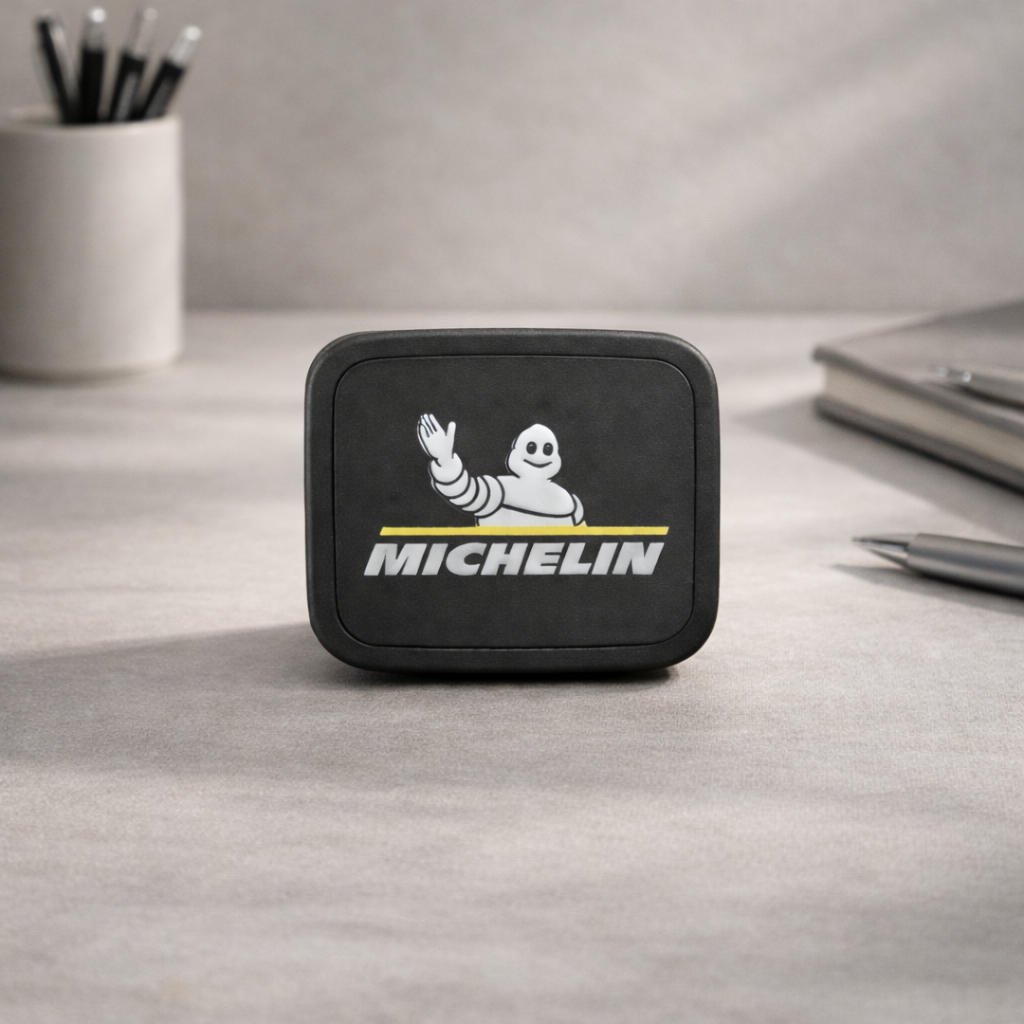 Un producto rectangular de color negro con la marca Michelin reposa sobre un escritorio gris, cerca de un portalápices y un cuaderno.