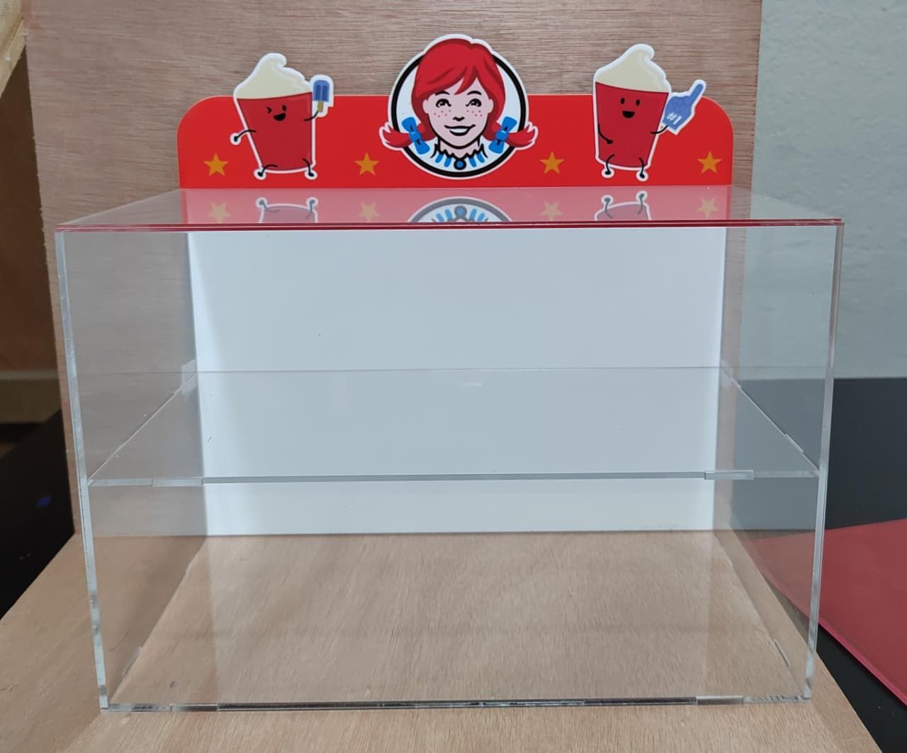 Una vitrina de dos niveles de acrílico transparente de Wendy's con un panel trasero rojo que muestra el logotipo de Wendy's y dos personajes mascota.