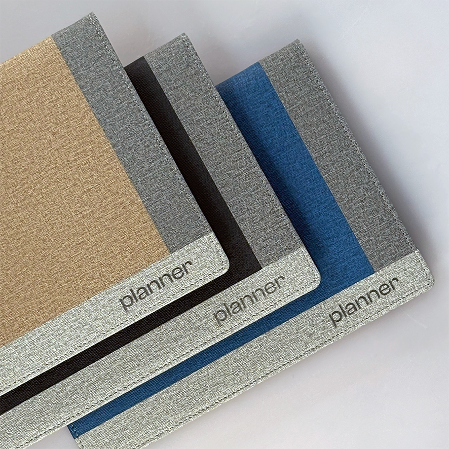 Tres agendas con cubiertas de tela en color dorado, gris y azul, cada una con una franja vertical en contraste y la palabra 