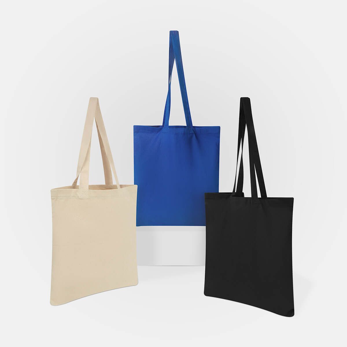 Tres bolsas de tela lisas de algodón en color beige, azul marino y negro, dispuestas sobre un fondo gris claro.