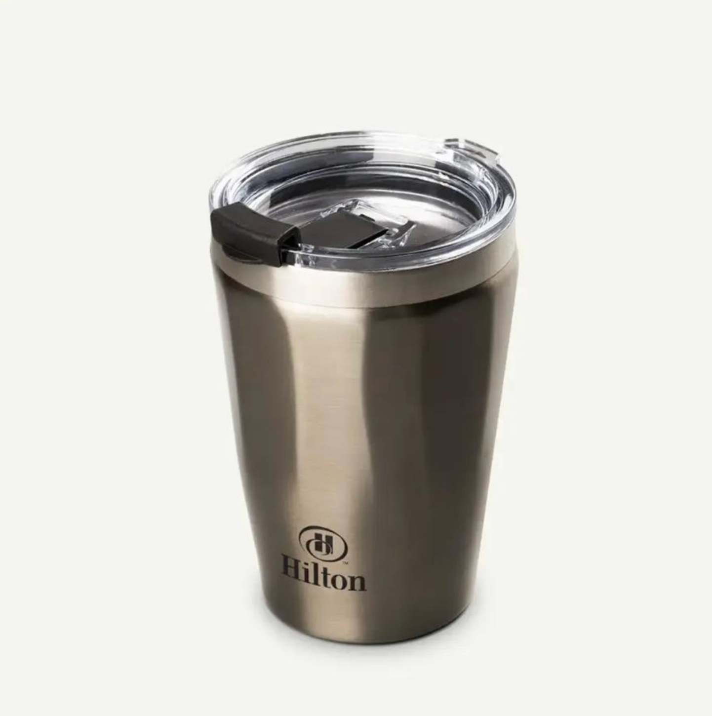 Un vaso térmico de viaje con la marca Hilton, de metal cepillado y con tapa de plástico transparente, sobre un fondo liso.