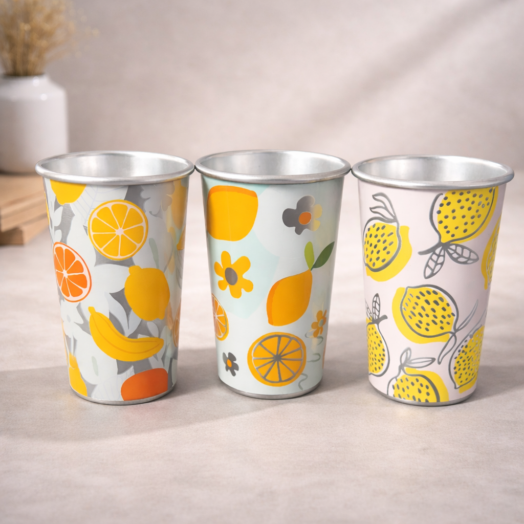 Tres vasos metálicos con motivos cítricos y frutales dispuestos sobre una superficie clara, con un jarrón al fondo.