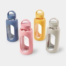 Cuatro botellas de agua de vidrio con fundas de silicona de colores azul, rosa, amarillo y beige, colocadas sobre un fondo blanco.