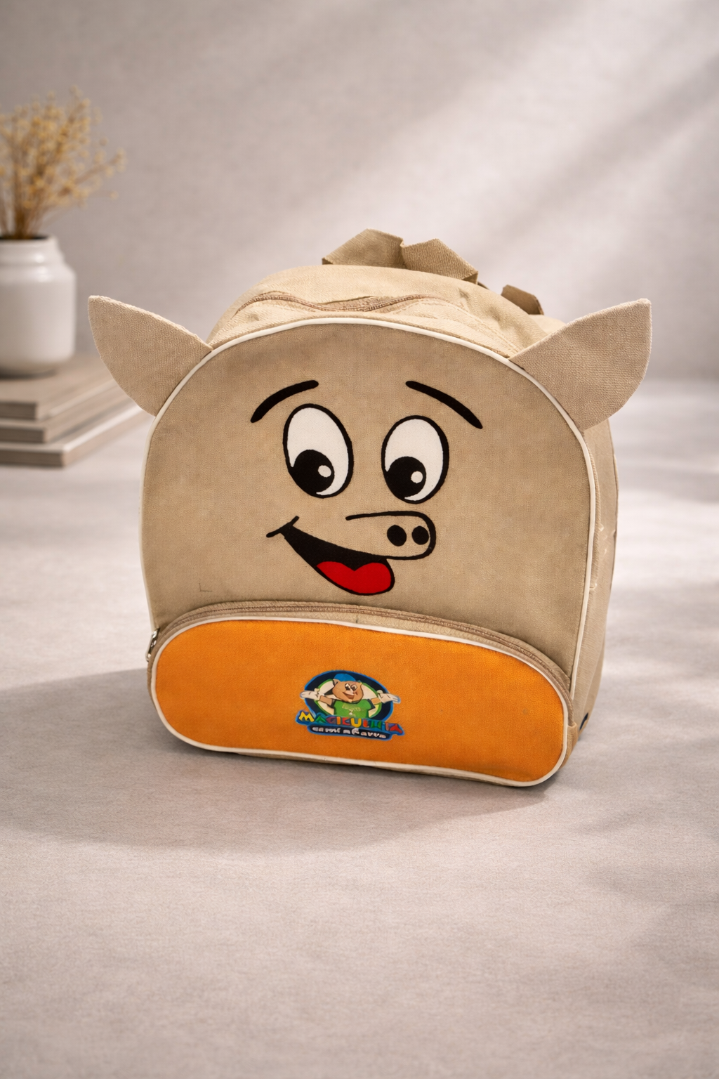 Una mochila beige con forma de cerdito de dibujos animados y un bolsillo inferior naranja, colocada sobre una superficie neutra junto a un jarrón.