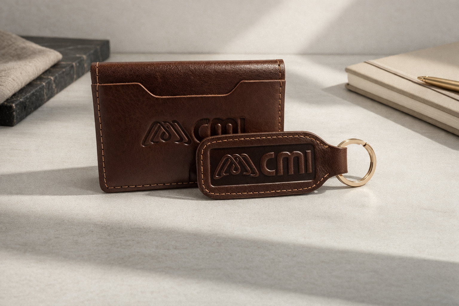 Un tarjetero de cuero marrón y un llavero a juego, ambos con el logotipo de CMI grabado en relieve, reposan sobre un escritorio blanco, con un bloc de notas cerca.