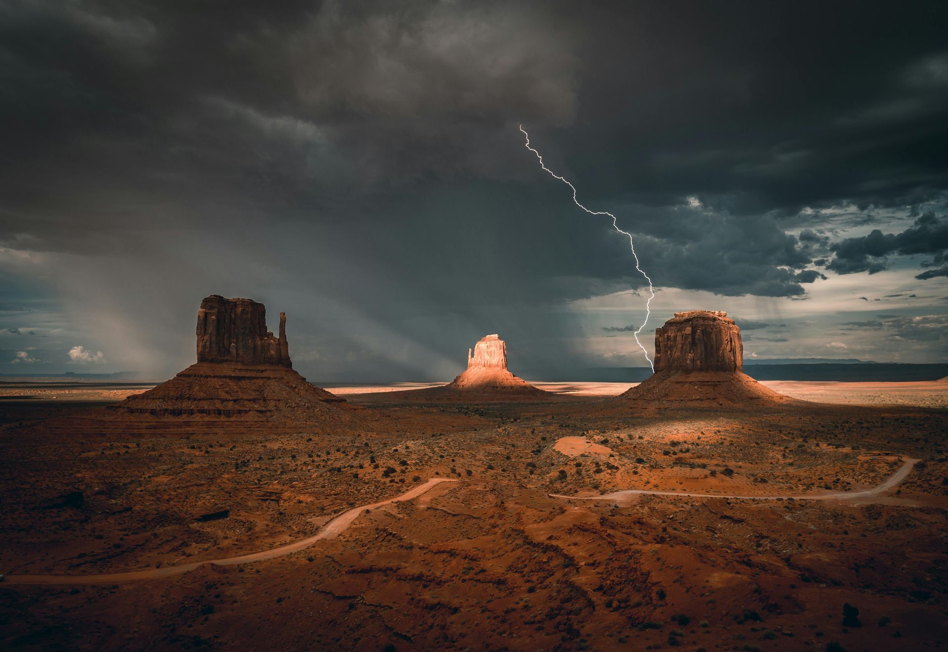 Un rayo cae cerca de tres mesetas de arenisca en Monument Valley bajo un cielo dramático, tormentoso y lleno de nubes.