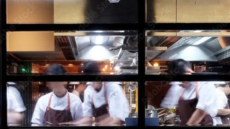 Chefs in white coats and aprons working in a busy kitchen with bright lights and stainless steel fixtures.