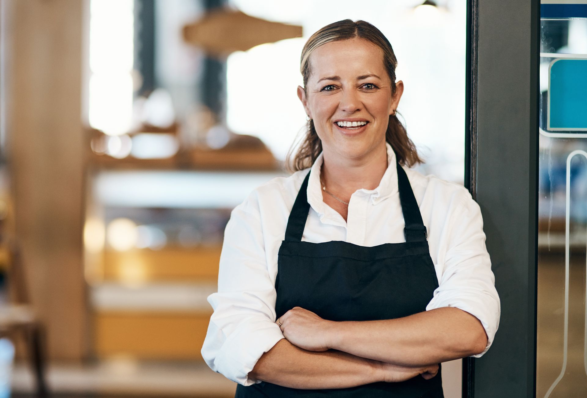 Man in apron smiles, arms crossed, behind a cafe counter. Bright lights, blurred background with other people.