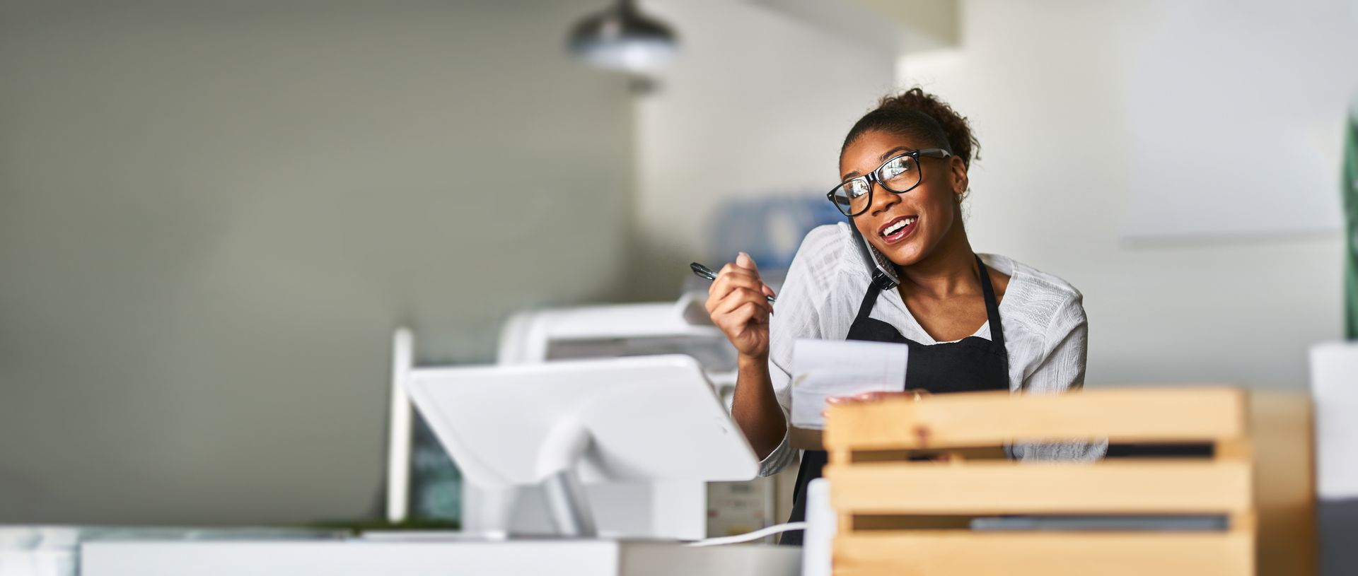 Person wearing glasses, apron, talking on phone, holding pen and paper behind a counter.