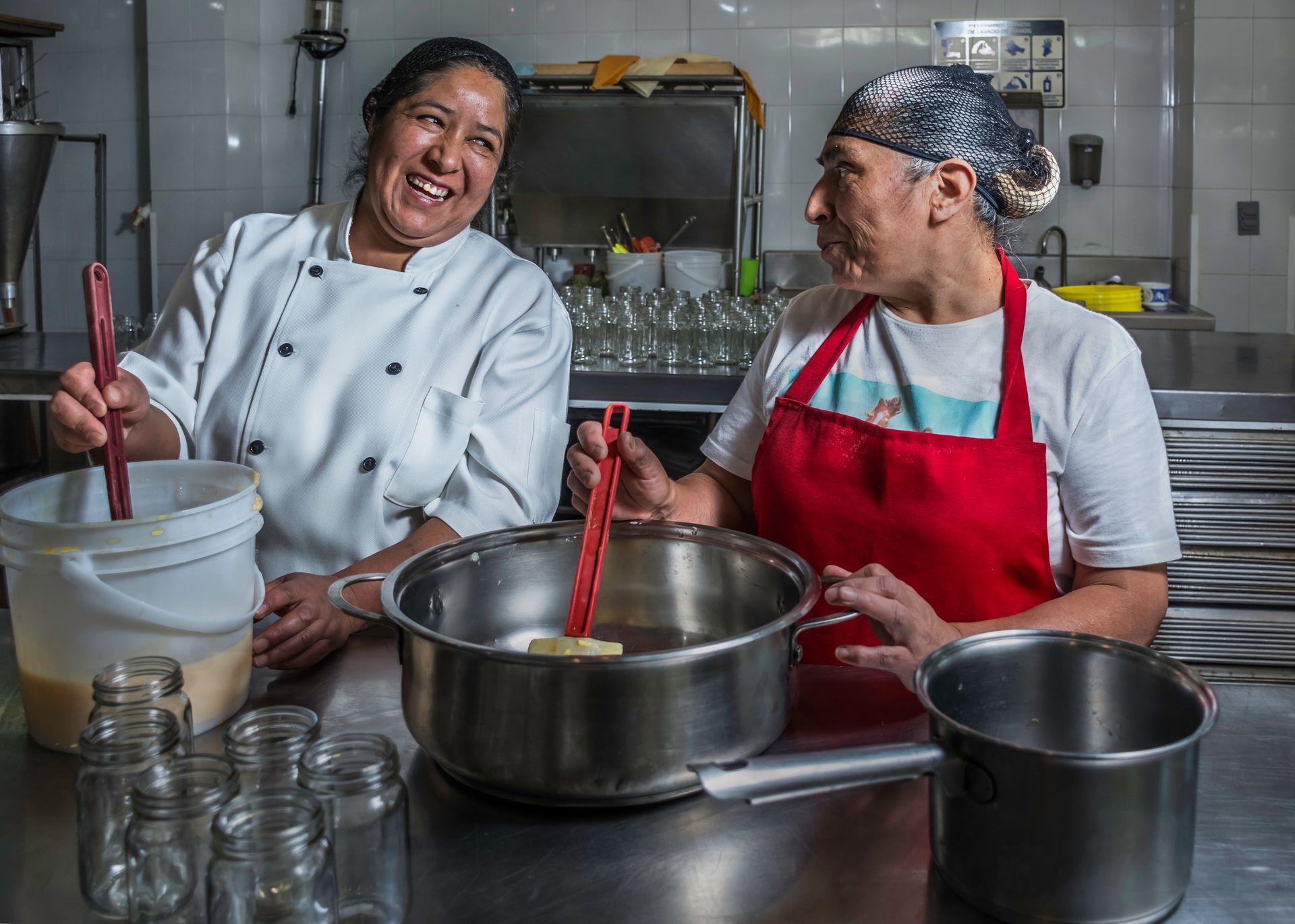 Two people cooking in a kitchen, one stirring a pot and the other smiling, with jars and other pots in the foreground.