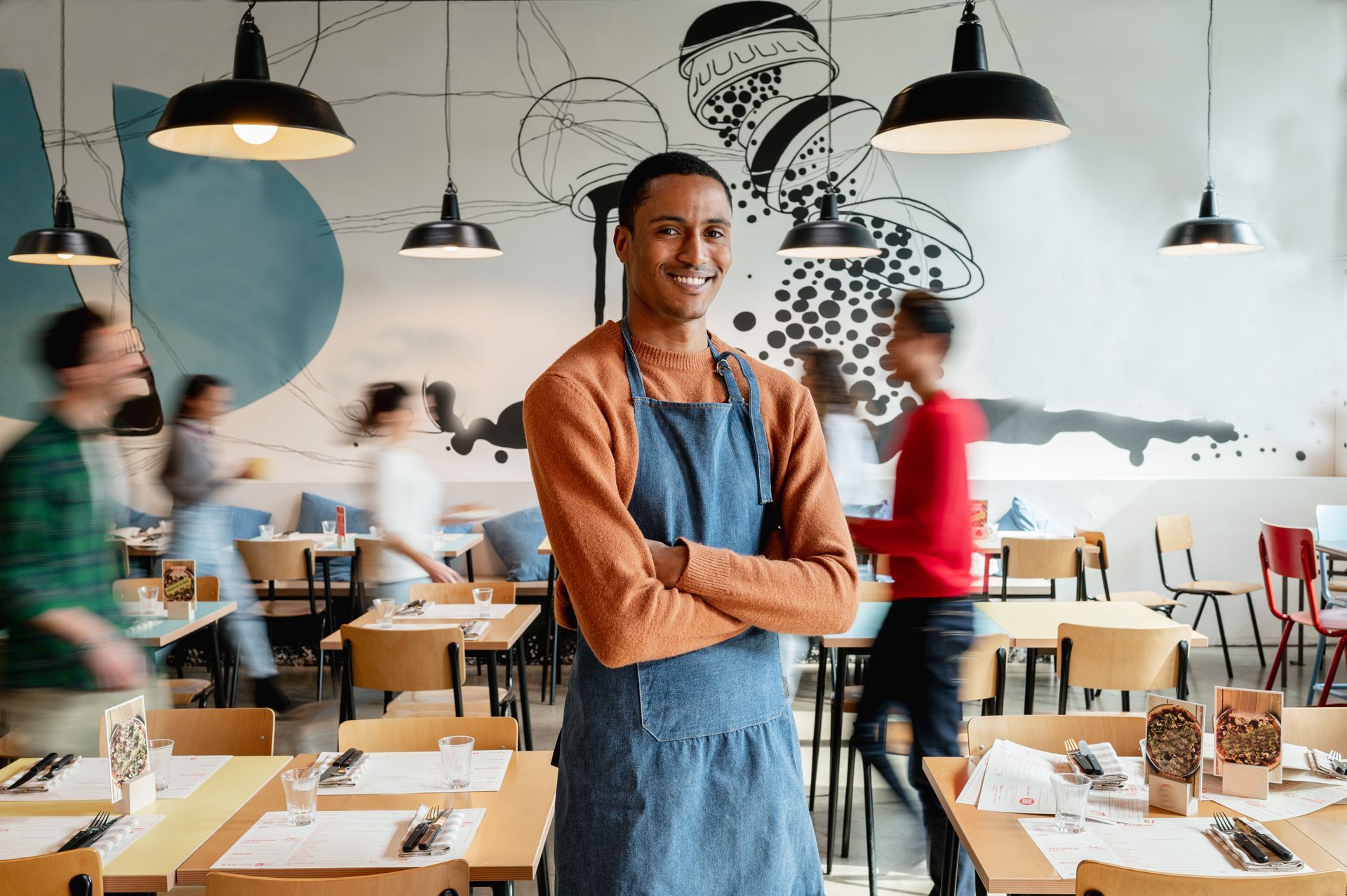 Man in apron smiles, arms crossed, behind a cafe counter. Bright lights, blurred background with other people.