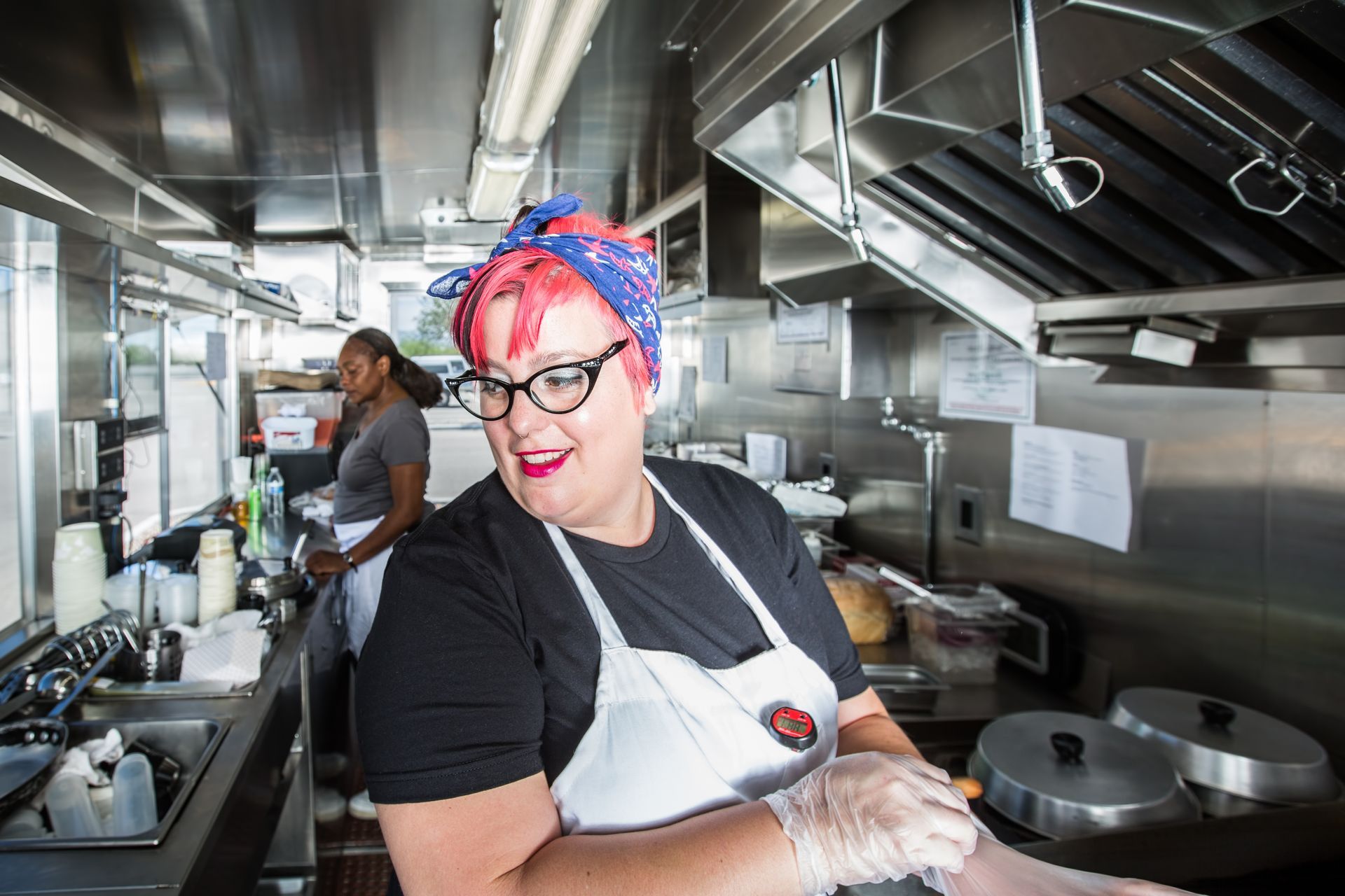 Woman in food truck kitchen smiles, wearing apron and gloves. Stainless steel counter and hood visible.