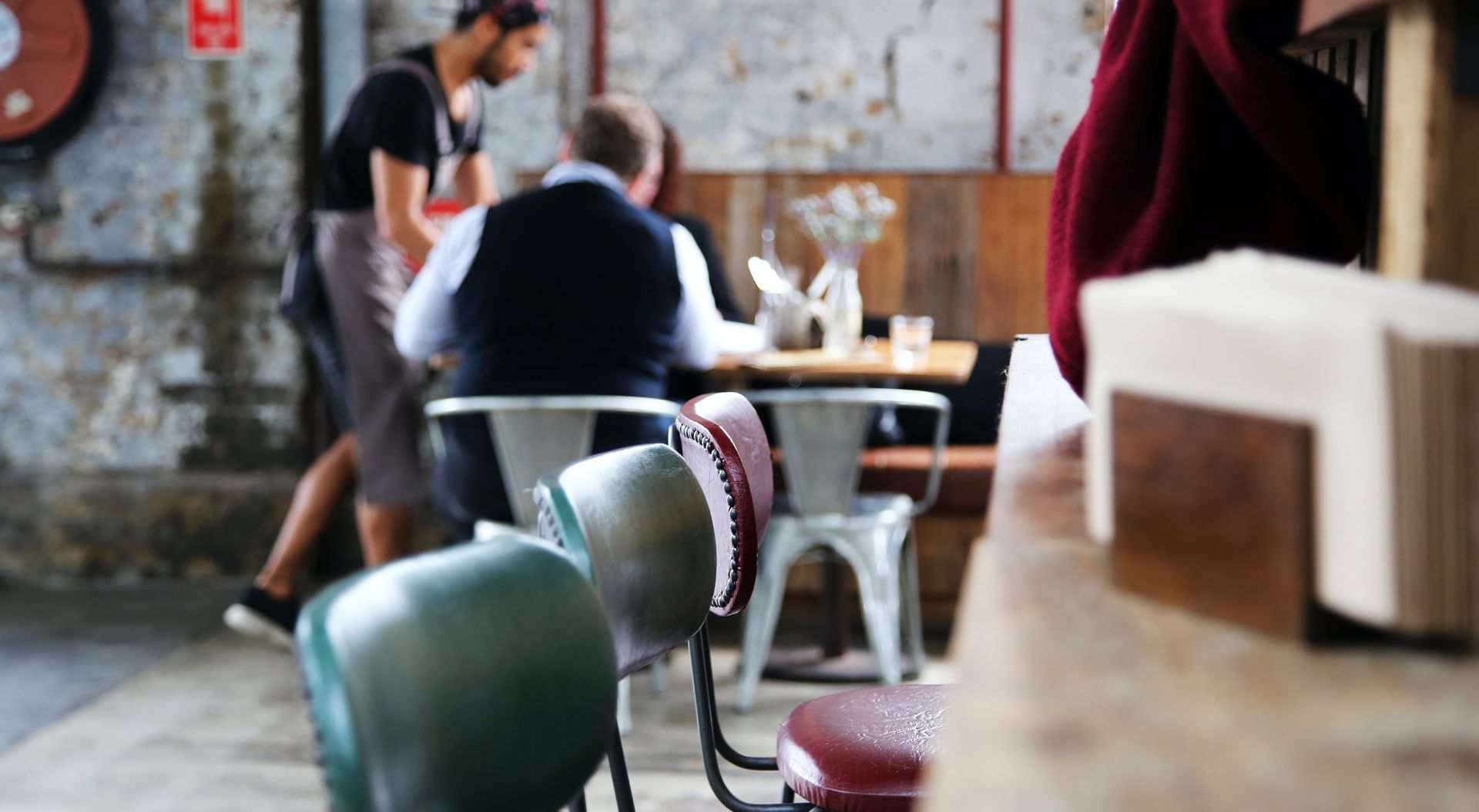 Cafe scene: waiter serves customers at a table with vintage chairs, rustic wall, and wooden counter in the foreground.