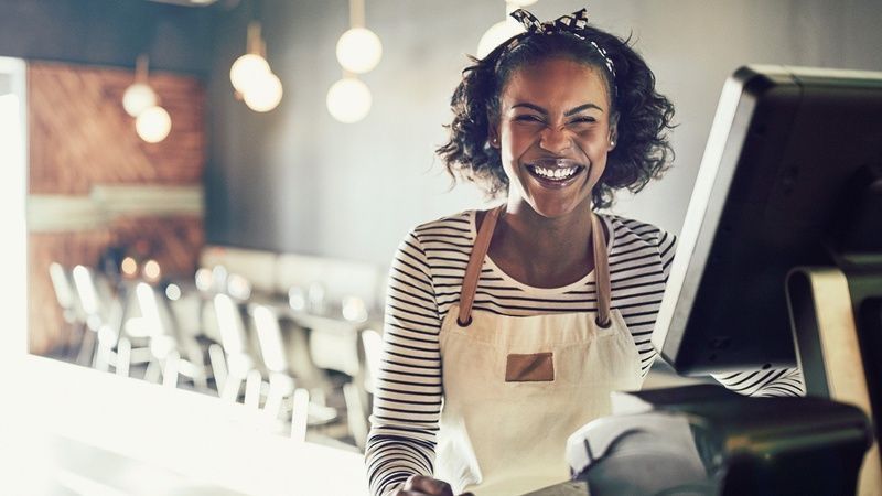 Woman with a big smile, wearing an apron, working at a POS system in a restaurant.