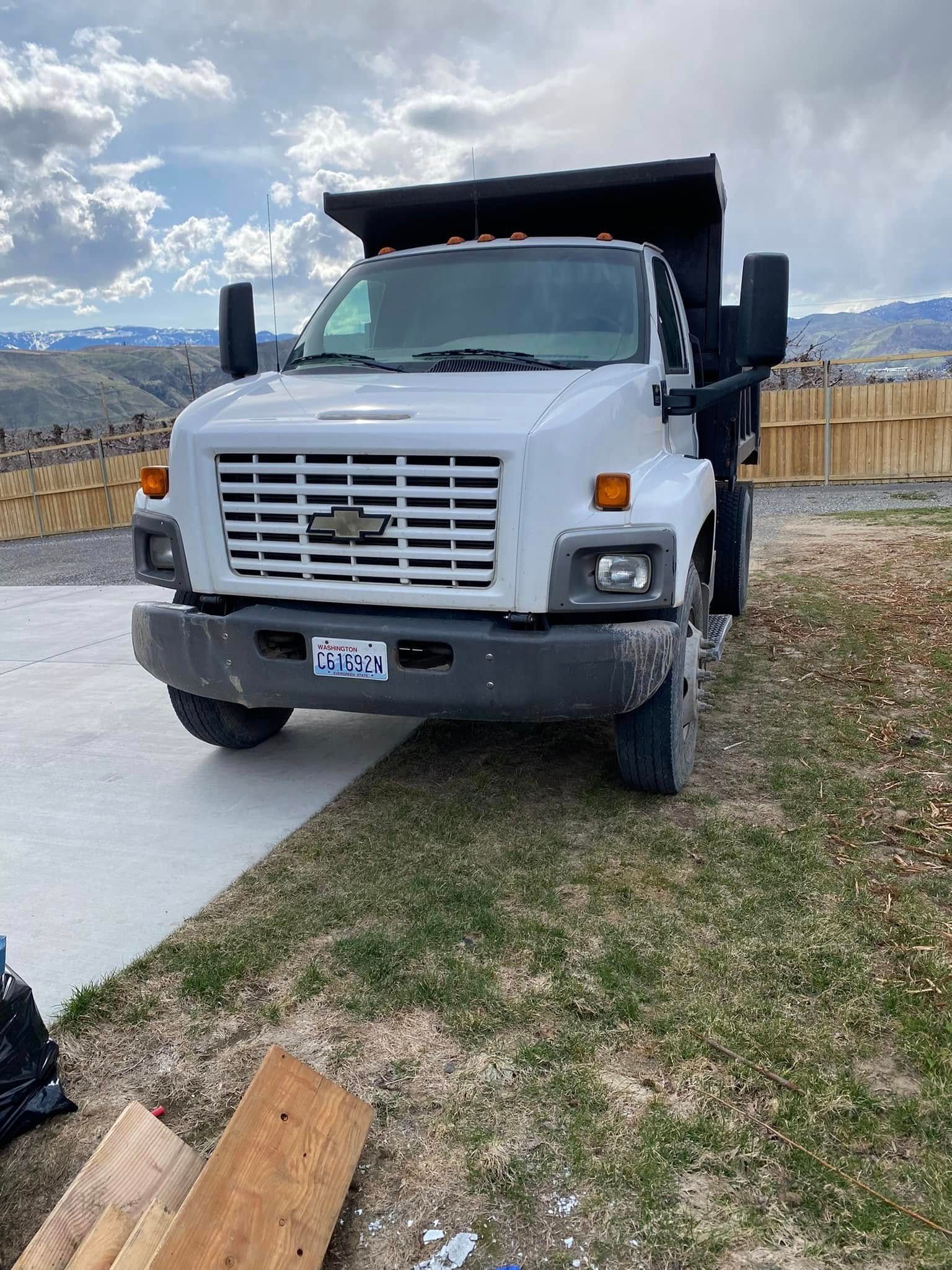 A white dump truck is parked in a driveway next to a pile of wood.