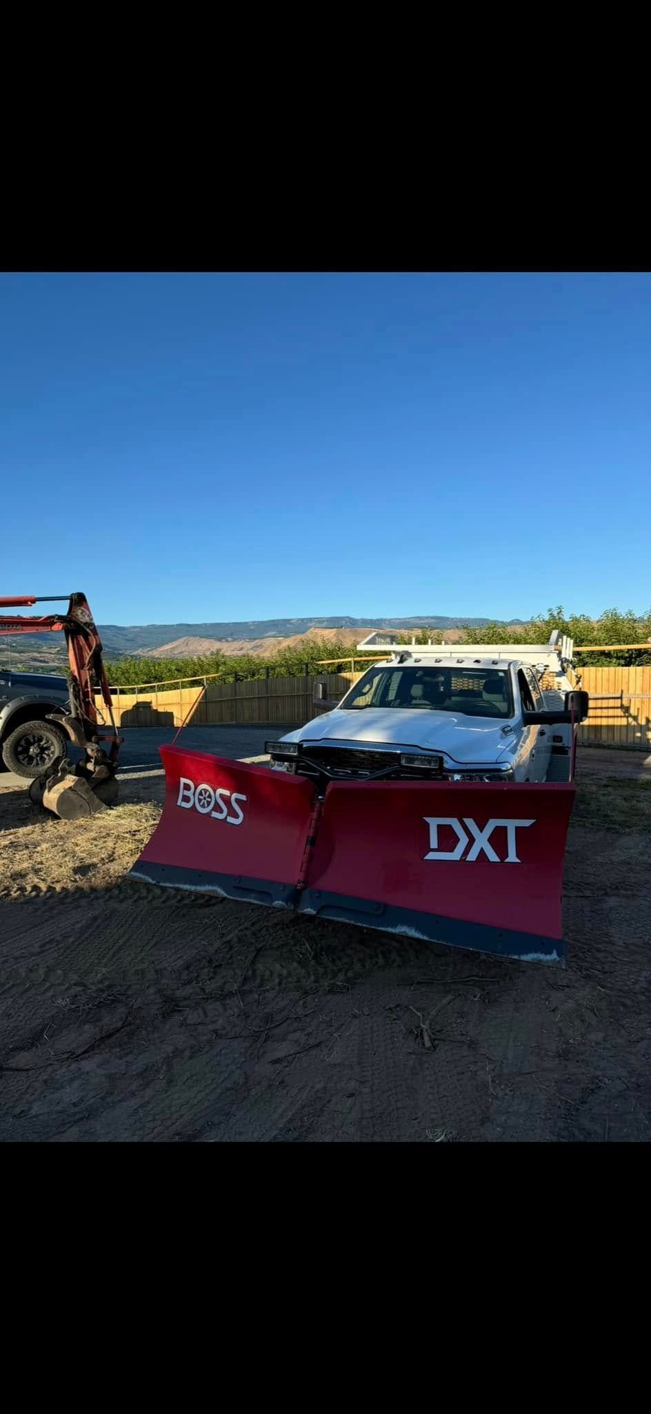 A snow plow is parked in a dirt lot next to a truck.