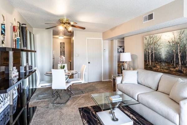 Cozy living room with cream sofa, glass coffee table, and a dining area. Artwork and trophies adorn the walls.