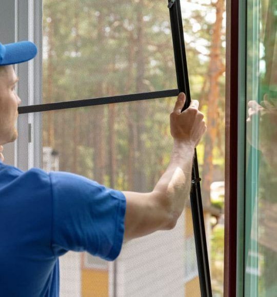A Man in a Blue Shirt is Adjusting a Screen on a Window — Budget Screens & Awnings in South Lismore, NSW