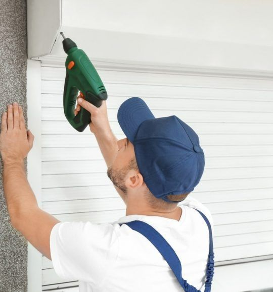 A Man in a Blue Hat is Using a Drill on a Wall — Budget Screens & Awnings in South Lismore, NSW