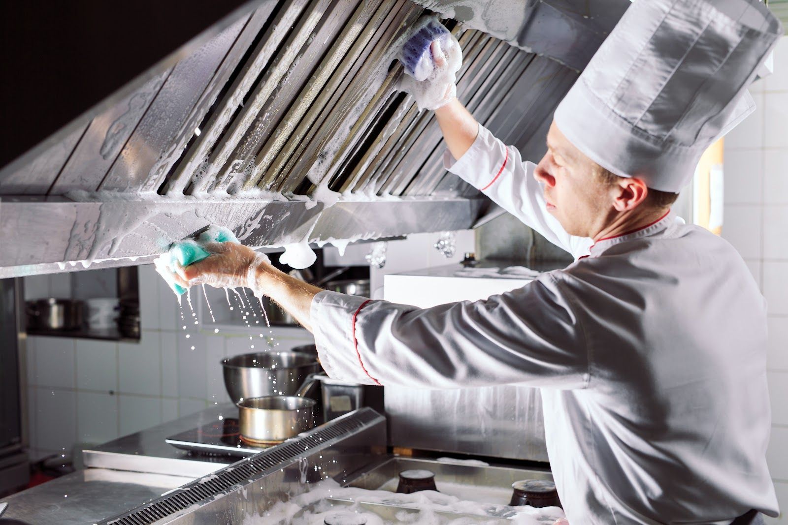 Chef cleaning a commercial kitchen hood with a sponge and soapy water.