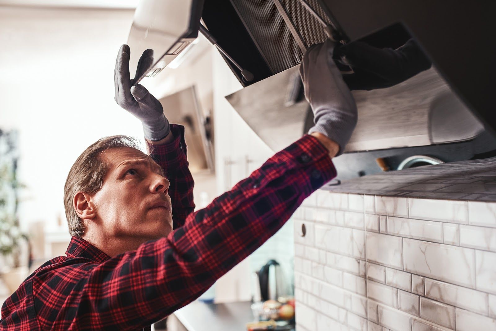 Man in red plaid shirt and gloves installing a range hood in a kitchen.
