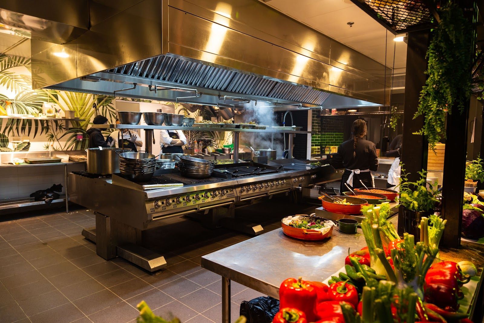 Restaurant kitchen with cooking equipment and chefs, fresh produce in foreground.
