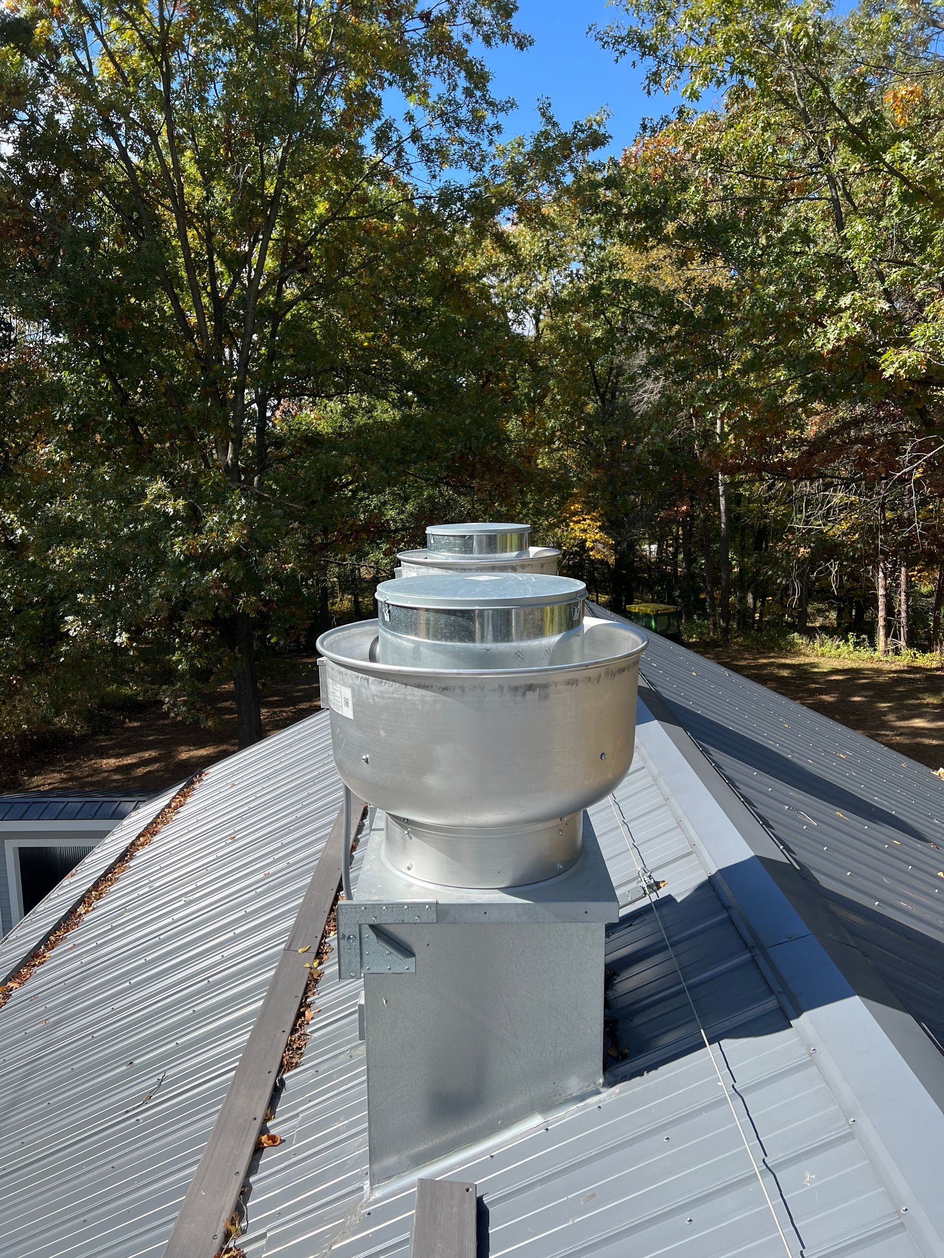 Metal exhaust vent on a corrugated metal roof, with trees in the background under a blue sky.