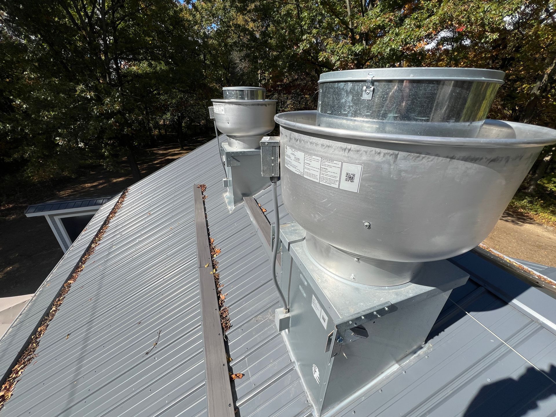 Two large metal roof exhaust vents on a gray metal roof, with trees in the background.