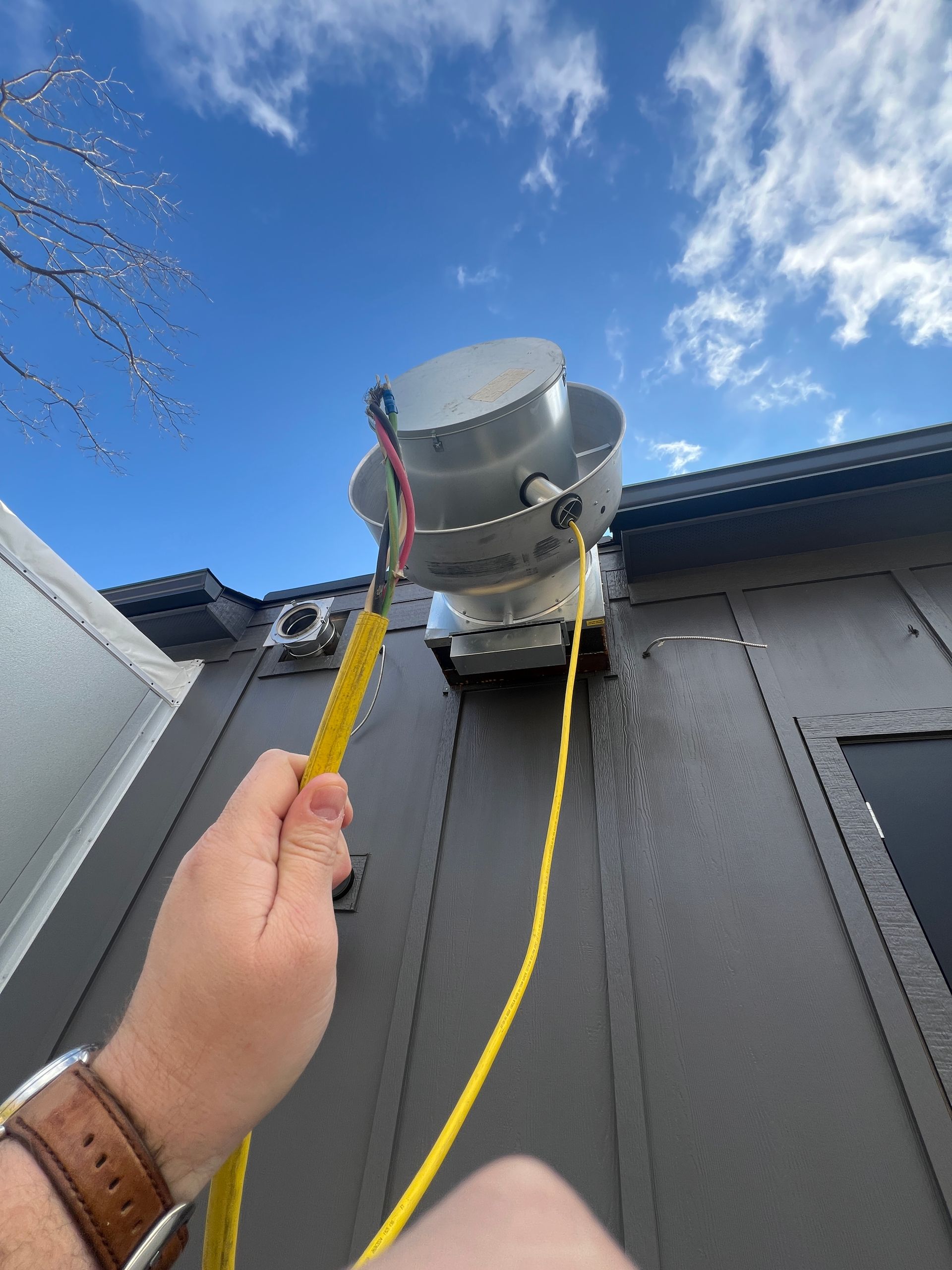 A person holding a yellow cable attached to a gray industrial device mounted on a building under a blue sky.