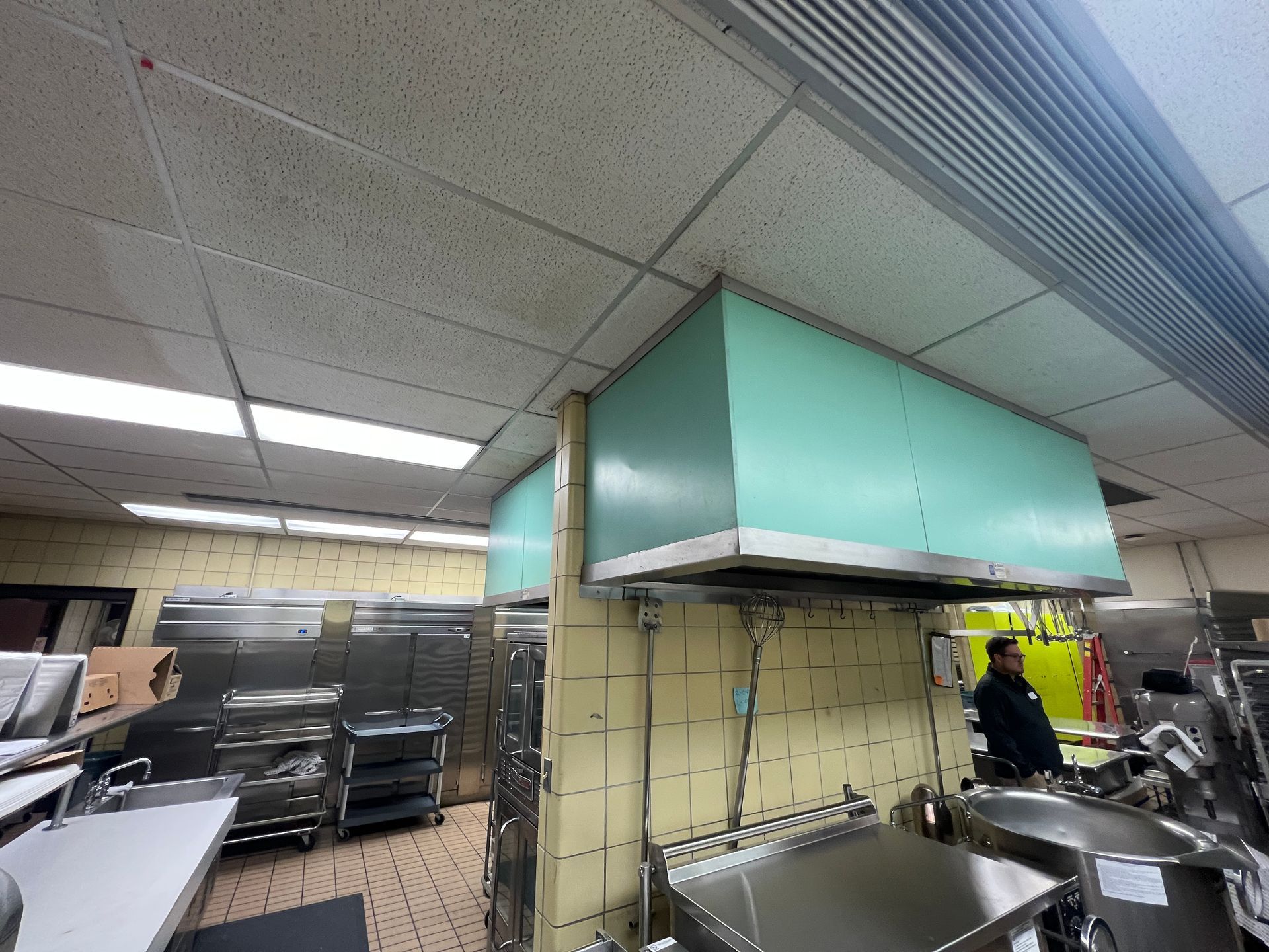 Commercial kitchen with teal exhaust hood, stainless steel appliances, and a person standing near equipment.