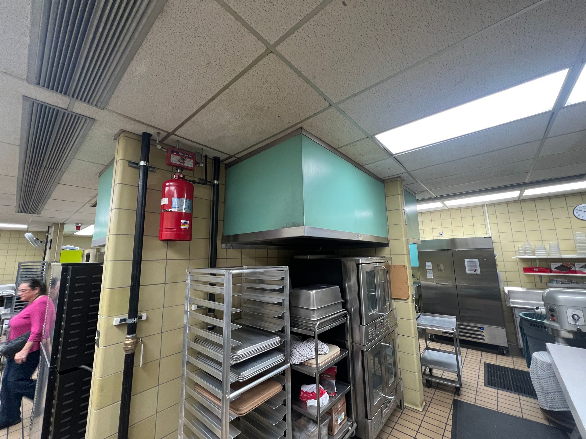 Kitchen interior with fire extinguisher, metal racks, and a person in pink shirt.