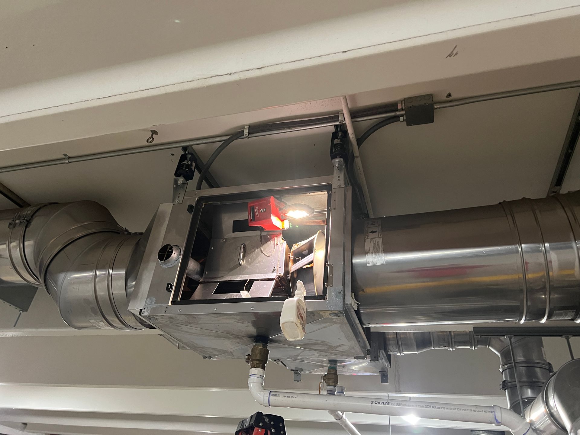 Metal HVAC equipment mounted to ceiling with an open access panel revealing internal components. Silver ductwork.