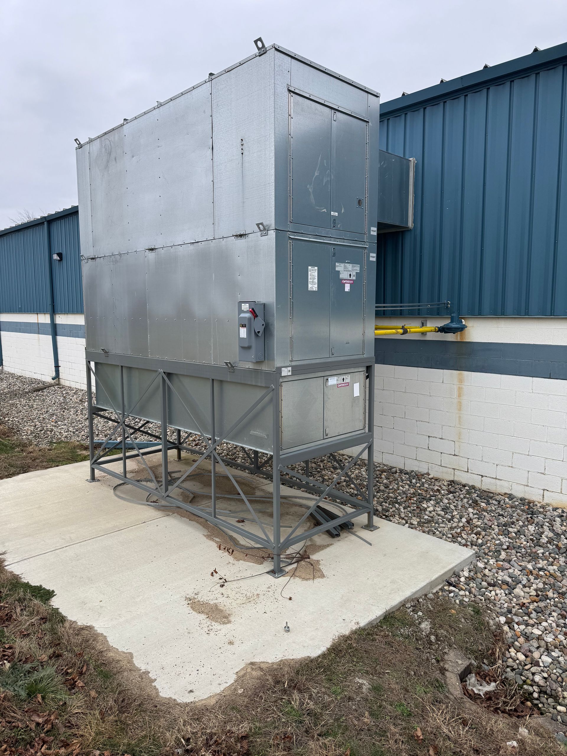 Large, metal industrial air handler unit, standing outdoors on a concrete pad near a blue building.