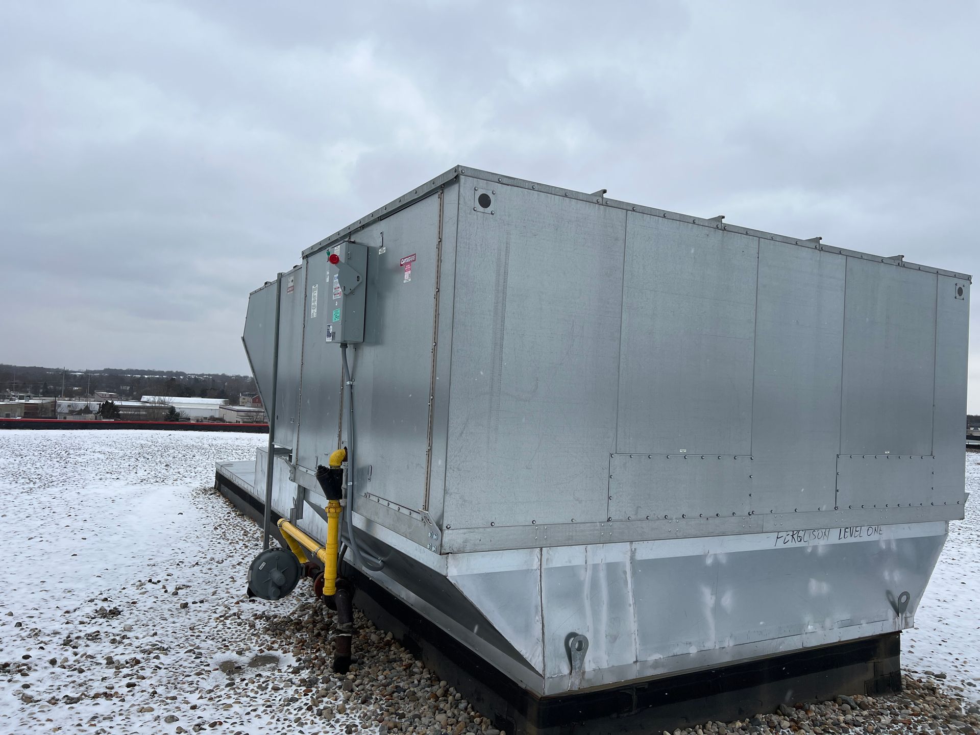 Rooftop HVAC unit on a building, gray metal with electrical box and yellow gas line. Snowy, overcast day.