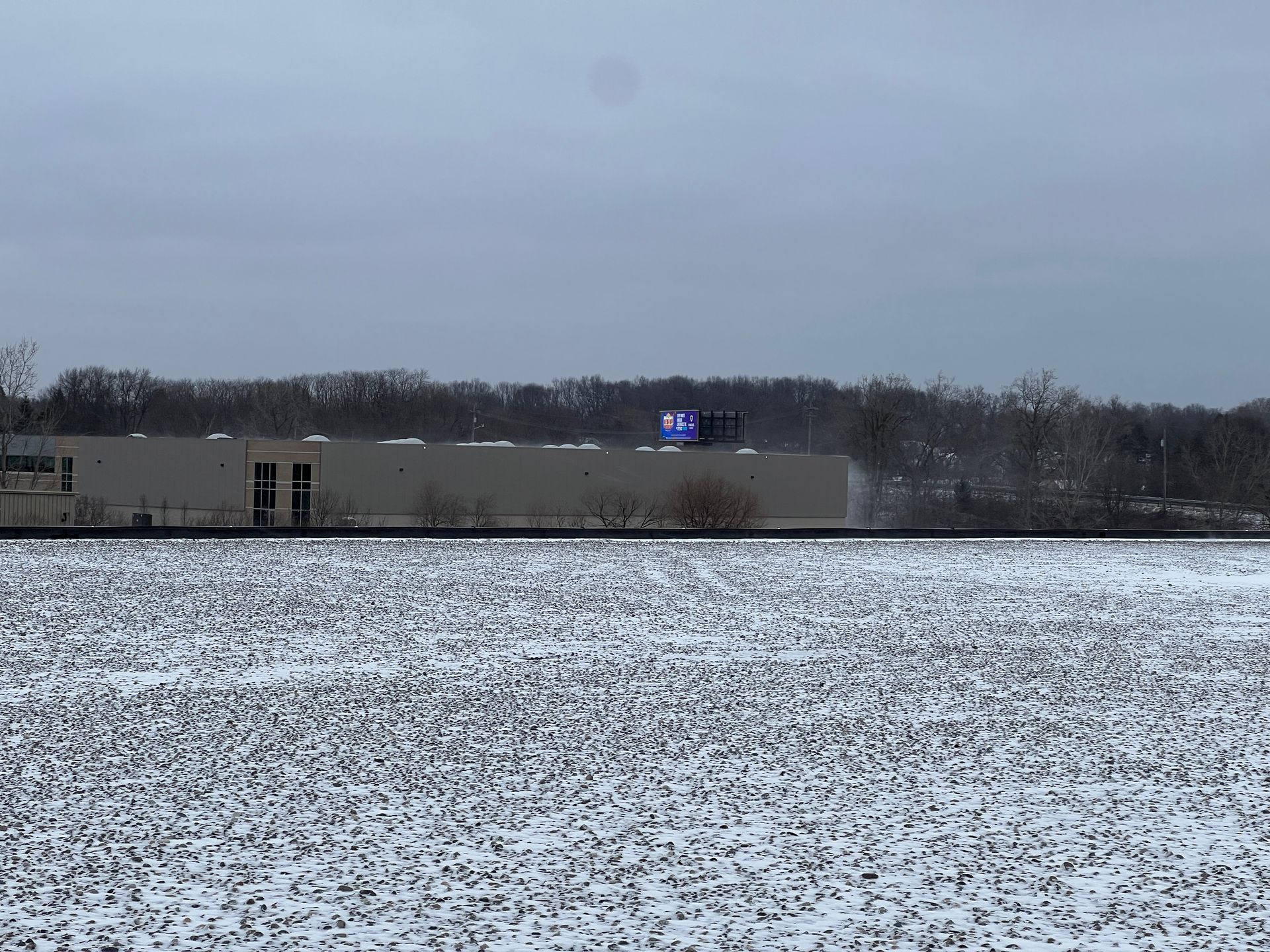 Snow-covered field with a long, white building in the background; a blue sign is on the roof. Overcast sky.