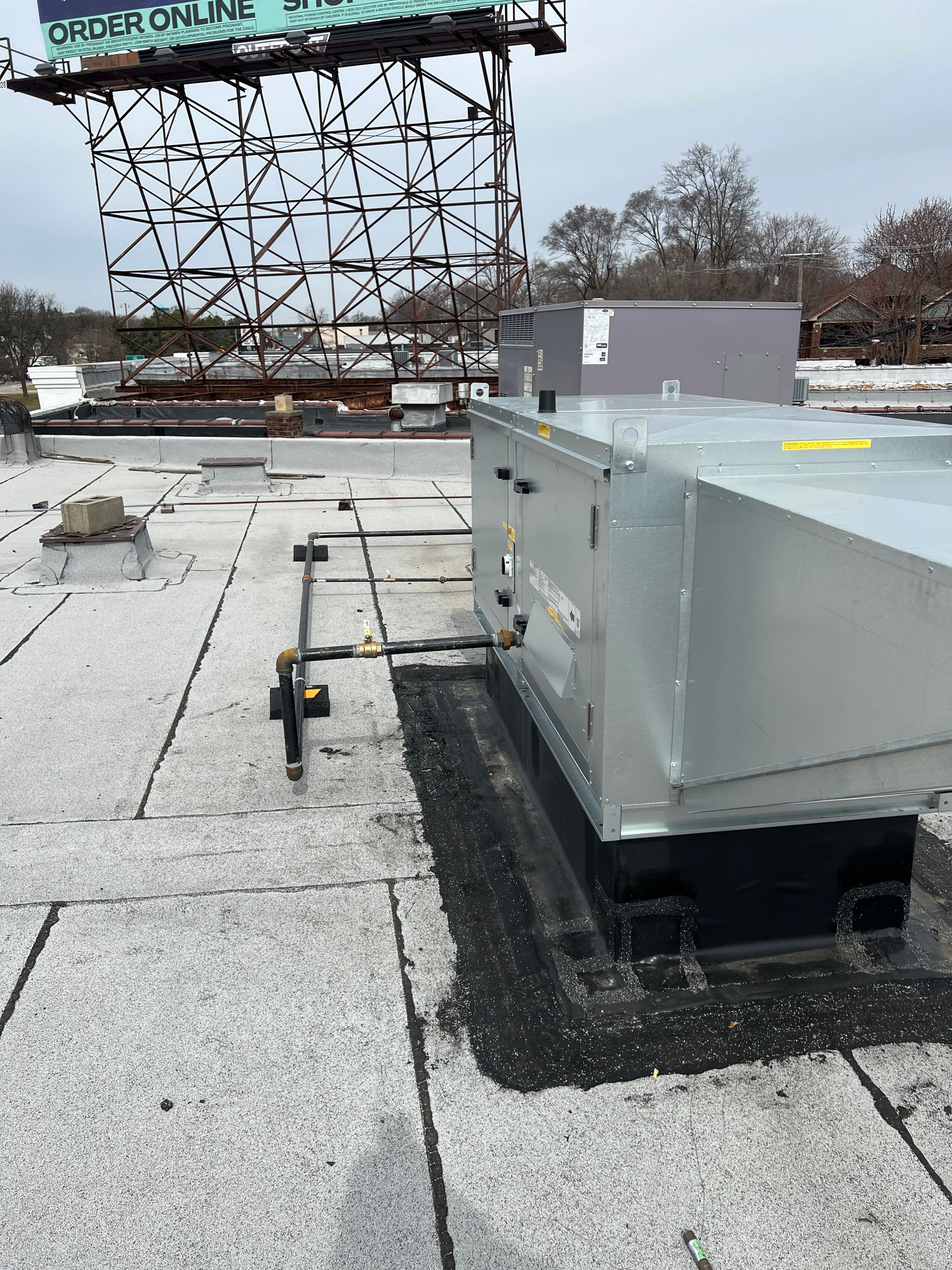 Rooftop HVAC unit on a flat roof. Metal ductwork and black sealant present, with a billboard in the background.
