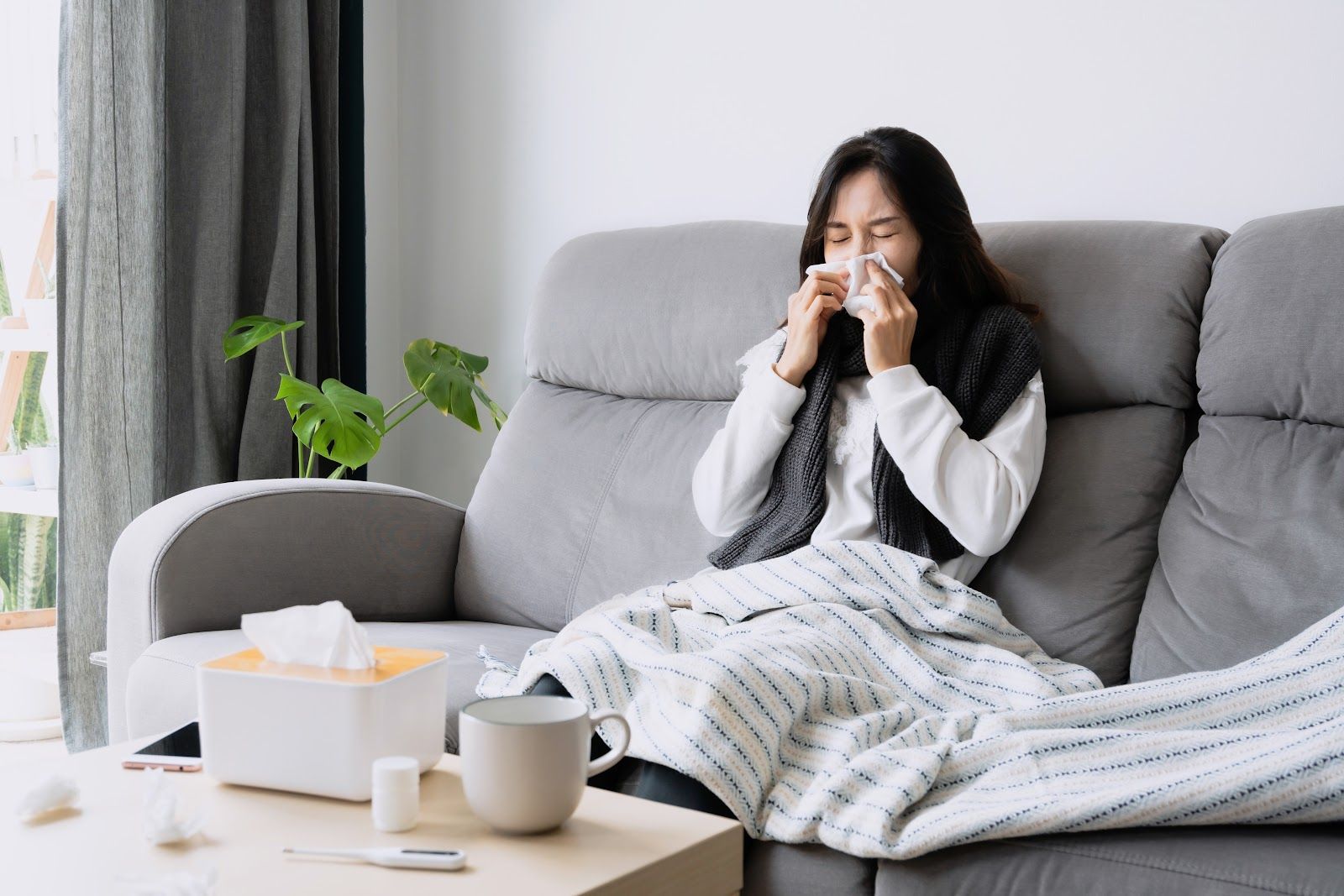 Woman on a couch blowing her nose with tissues, appears to have a cold.