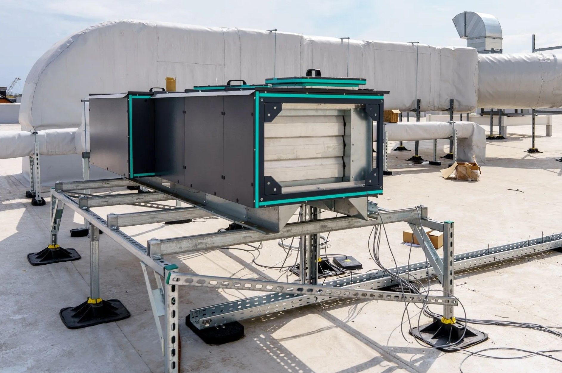 HVAC equipment on a rooftop, supported by a metal frame. Black box with teal accents, connected to ducts.